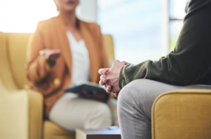 Two people in a counseling session: a therapist sits in the background with a tablet while the client sits in the foreground, hands clasped in the chair.