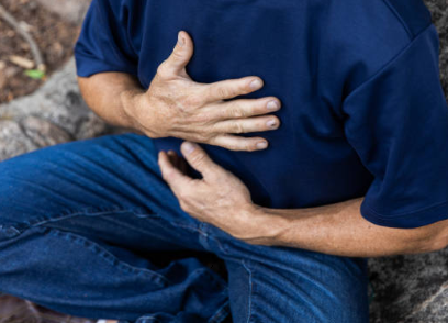 Person in a blue shirt sits outdoors, clutching their midsection with one hand and resting the other on their chest, suggesting abdominal or chest pain.